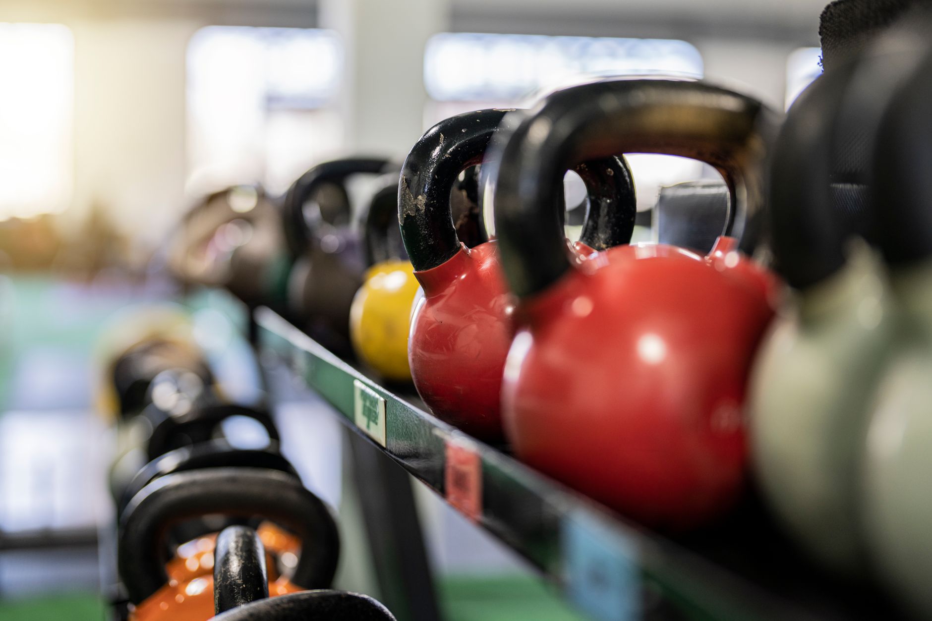 Fileira de kettlebells sobre uma prateleira de aço, em primeiro plano, com o salão de treino desfocado atrás.